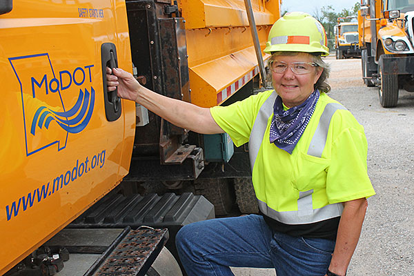 lady in yellow hard hat and vest grasping door of large dump truck