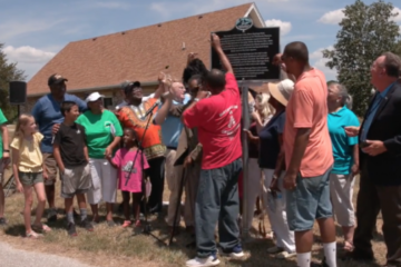 small diverse crowd watches unveiling of trail marker sign